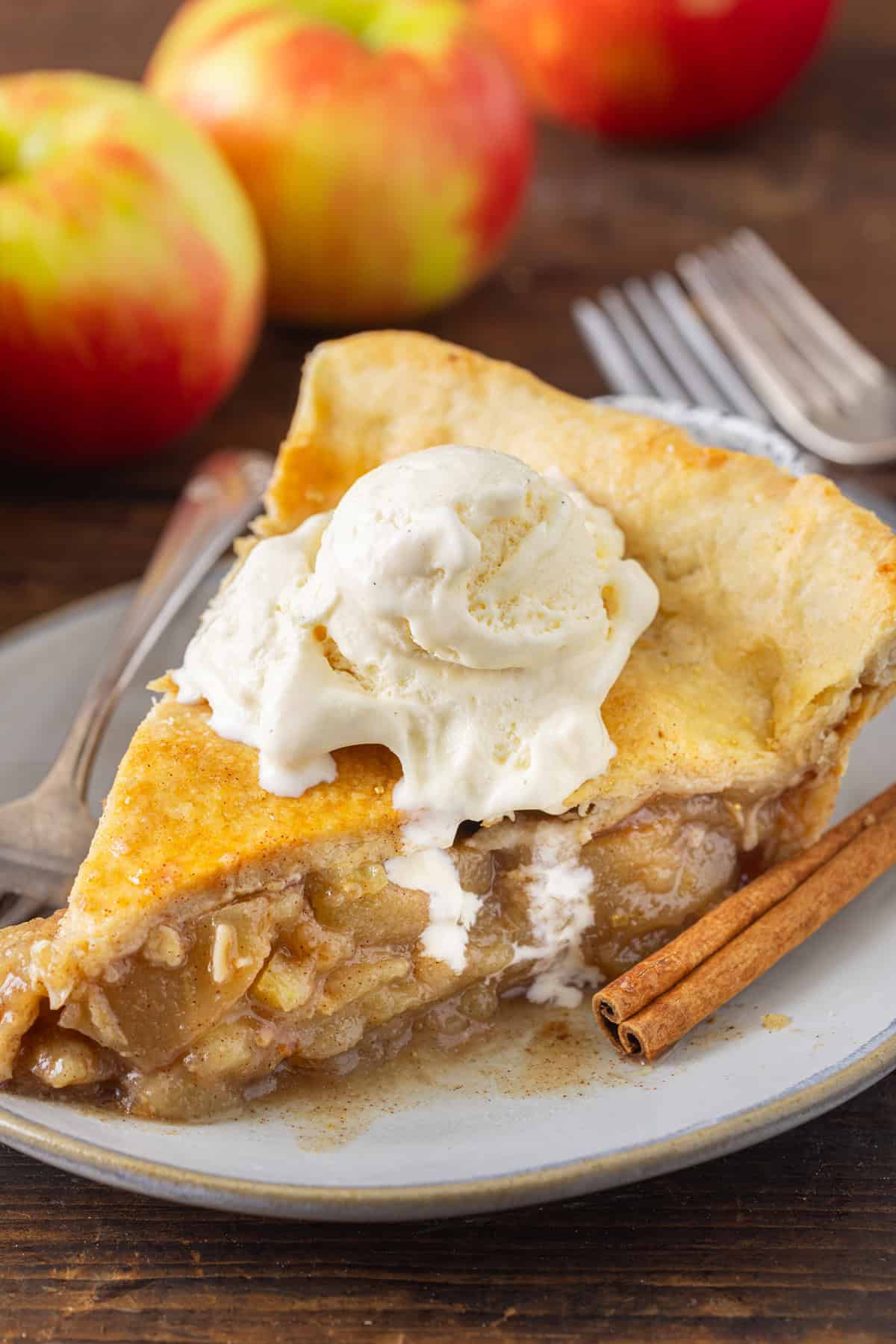 A slice of warm apple pie recipe topped with melting vanilla ice cream, served on a plate with a cinnamon stick and fresh apples in the background.