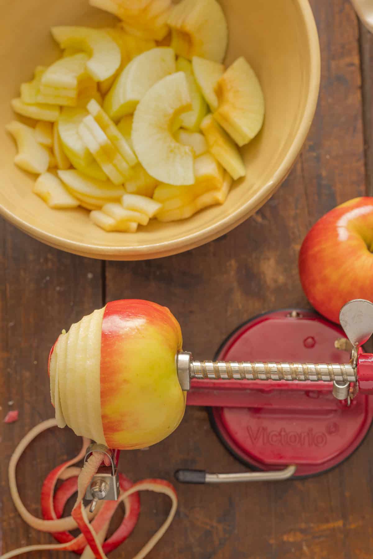An apple peeler in action, peeling a red and yellow apple while a bowl of freshly peeled and sliced apples sits nearby on a rustic wooden table.