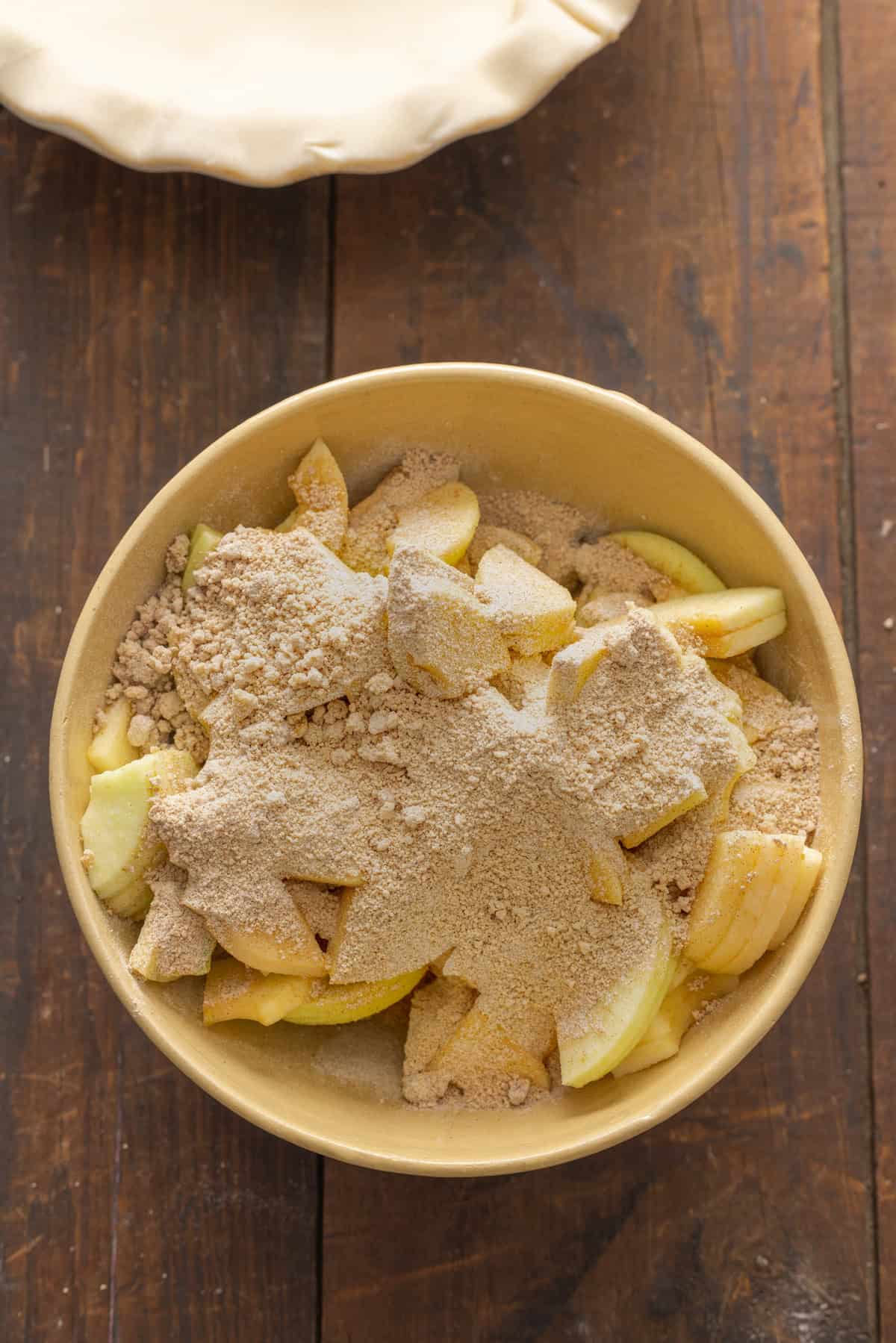 Apple slices coated with cinnamon and brown sugar in a bowl, ready to be mixed, with a pie crust visible in the background.