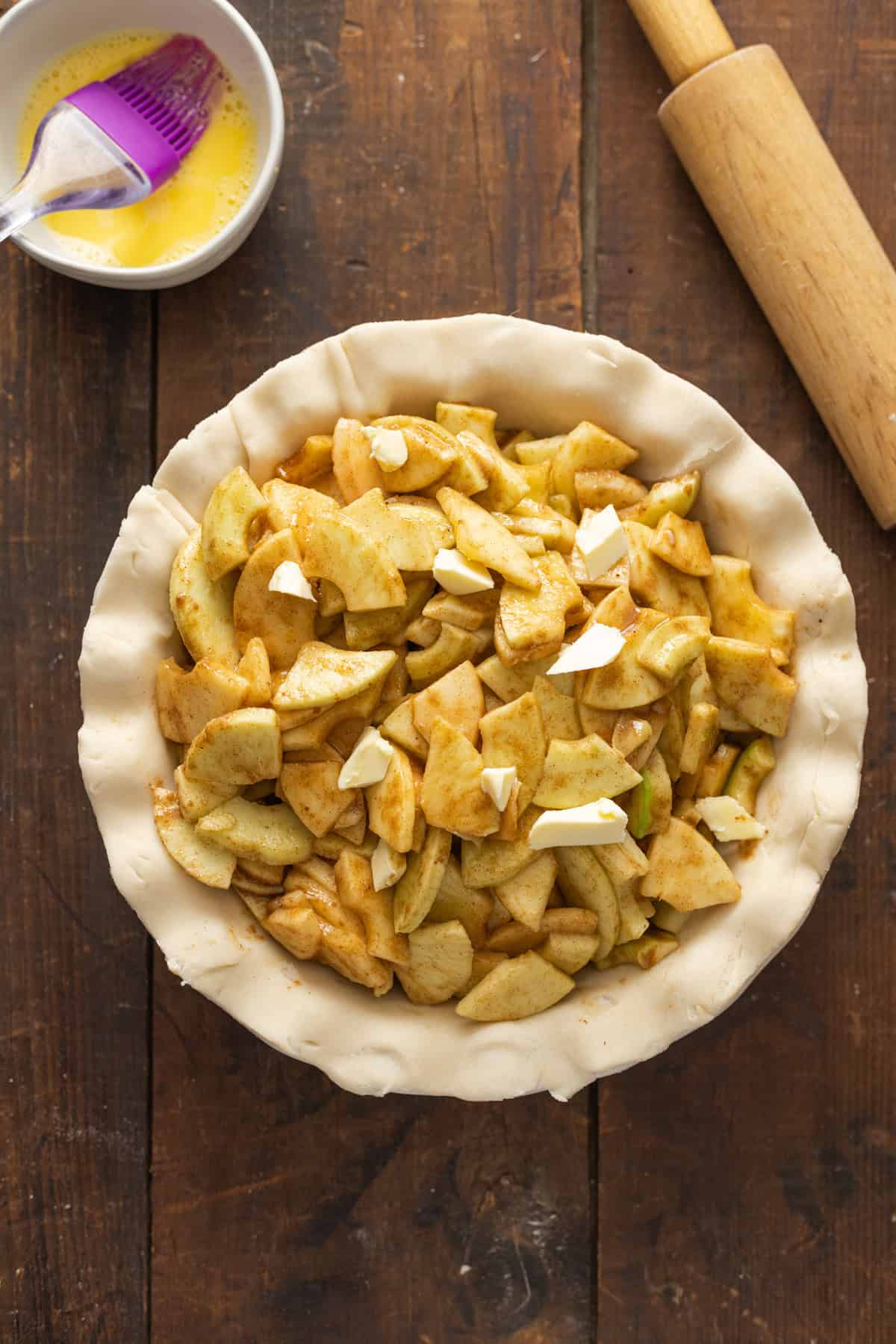 The top view of a pie crust filled with cinnamon-coated apple slices and small pats of butter, ready to be covered with the top crust.
