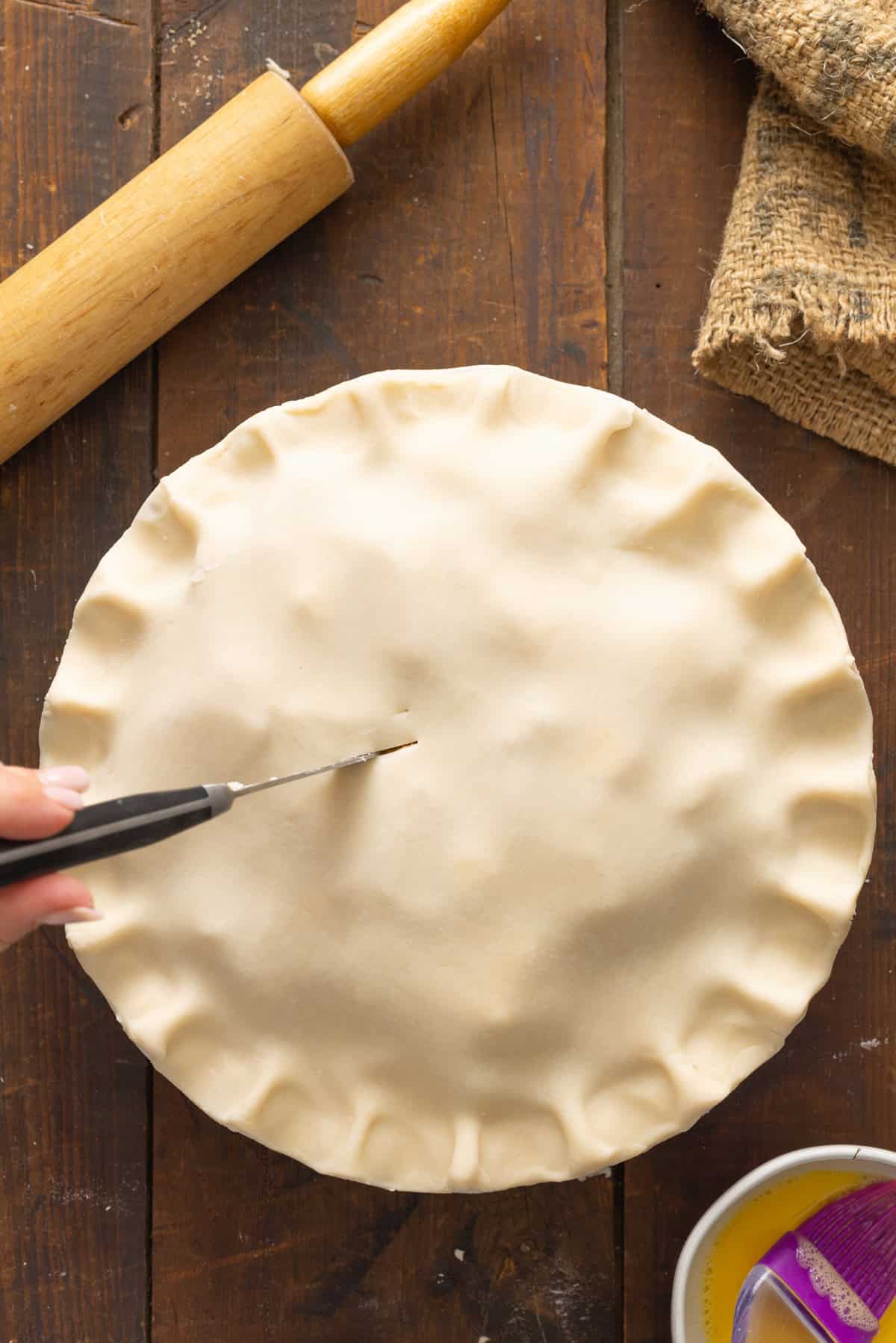 A hand cutting slits into the top crust of an unbaked apple pie recipe, ready for the oven, with a rolling pin and a bowl of egg wash beside it.