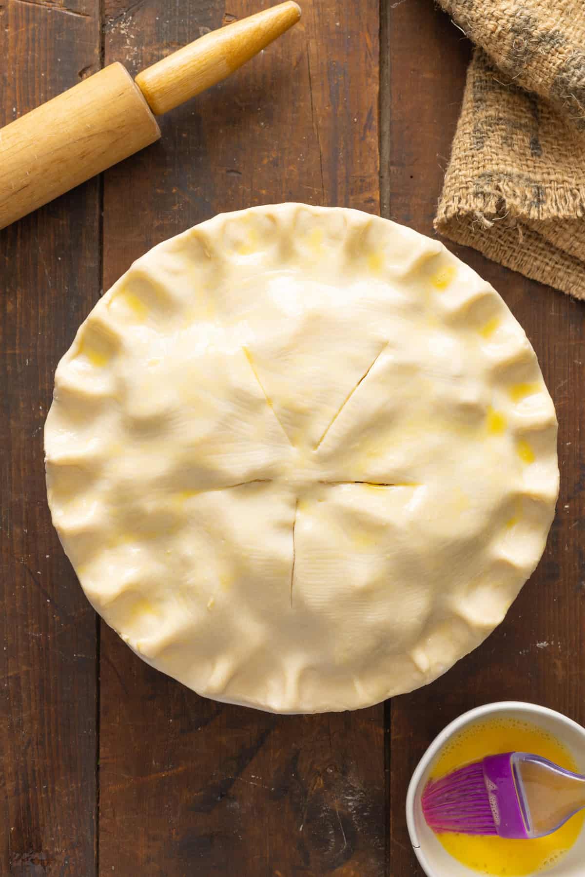An unbaked apple pie recipe with a golden egg wash applied on top, sitting on a wooden surface next to a rolling pin and a bowl of egg wash.