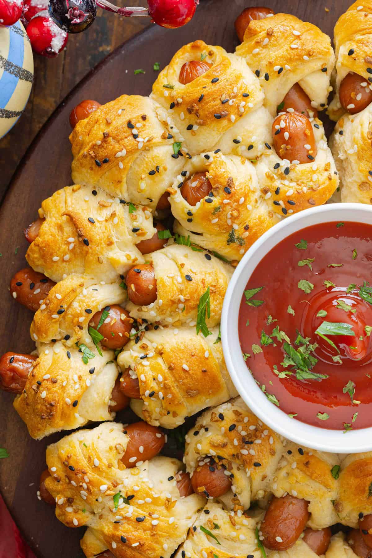 A full Pigs in a Blanket wreath displayed on a wooden board, garnished with rosemary and sesame seeds, with holiday ornaments in the background.