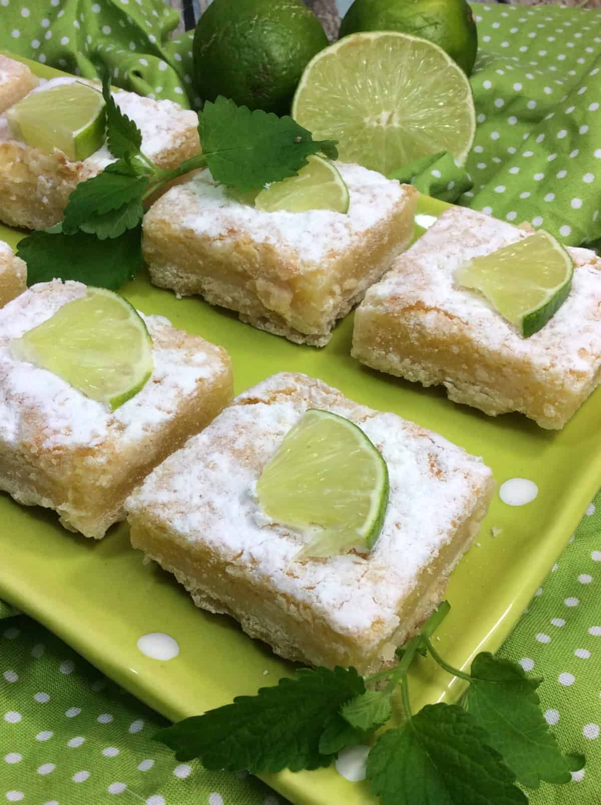 Key lime bars arranged on a green plate, topped with powdered sugar and lime wedges, surrounded by limes and green fabric