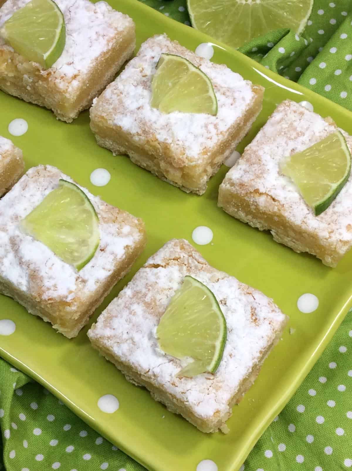 Close-up of key lime bars topped with powdered sugar and a fresh lime wedge on a green polka-dotted plate.