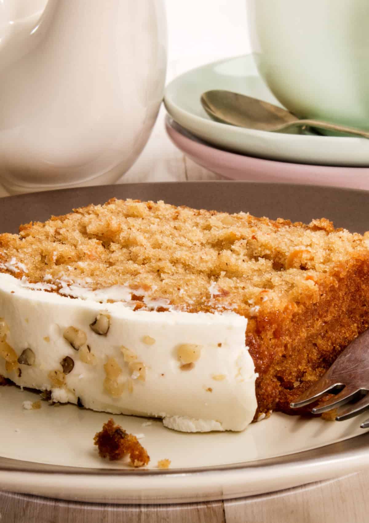 This Carrot Cake Loaf is perfectly moist and tender in the middle, bursting with warm spiced flavors and plenty of fresh carrot goodness. Topped with a rich and tangy cream cheese frosting, every slice is pure comfort and absolutely irresistible! A plated slice of carrot cake with a thick layer of cream cheese frosting, garnished with nuts, served with tea or coffee.