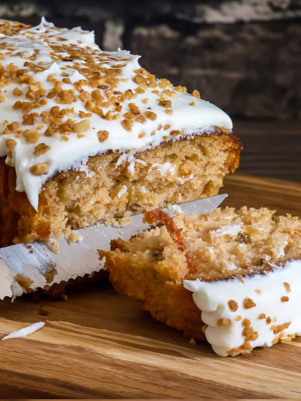 This Carrot Cake Loaf is perfectly moist and tender in the middle, bursting with warm spiced flavors and plenty of fresh carrot goodness. Topped with a rich and tangy cream cheese frosting, every slice is pure comfort and absolutely irresistible! A close-up of a knife cutting into a frosted carrot cake loaf, revealing its soft and moist texture with specks of shredded carrot.