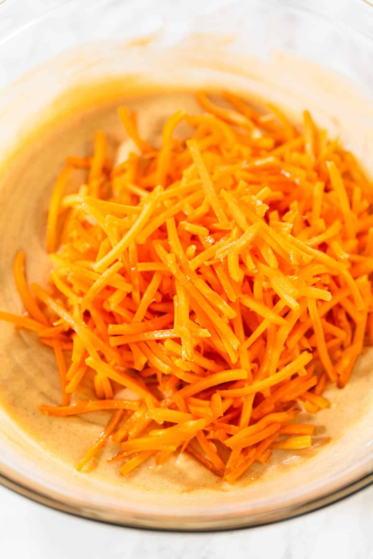 This Carrot Cake Loaf is perfectly moist and tender in the middle, bursting with warm spiced flavors and plenty of fresh carrot goodness. Topped with a rich and tangy cream cheese frosting, every slice is pure comfort and absolutely irresistible! A glass bowl containing a heap of freshly shredded carrots sitting on top of a spiced cake batter.