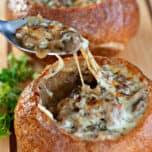 Two bread bowls filled with Philly Cheesesteak Soup, with golden, bubbly melted cheese on top, placed on a wooden cutting board with a spoon and fresh parsley for garnish.
