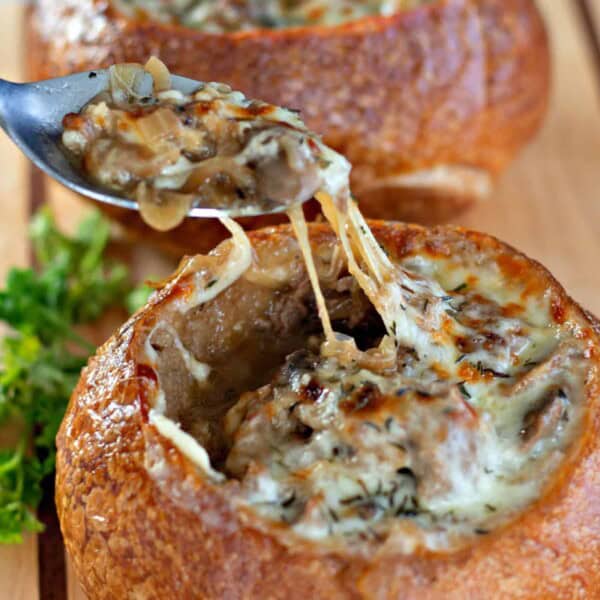 Two bread bowls filled with Philly Cheesesteak Soup, with golden, bubbly melted cheese on top, placed on a wooden cutting board with a spoon and fresh parsley for garnish.