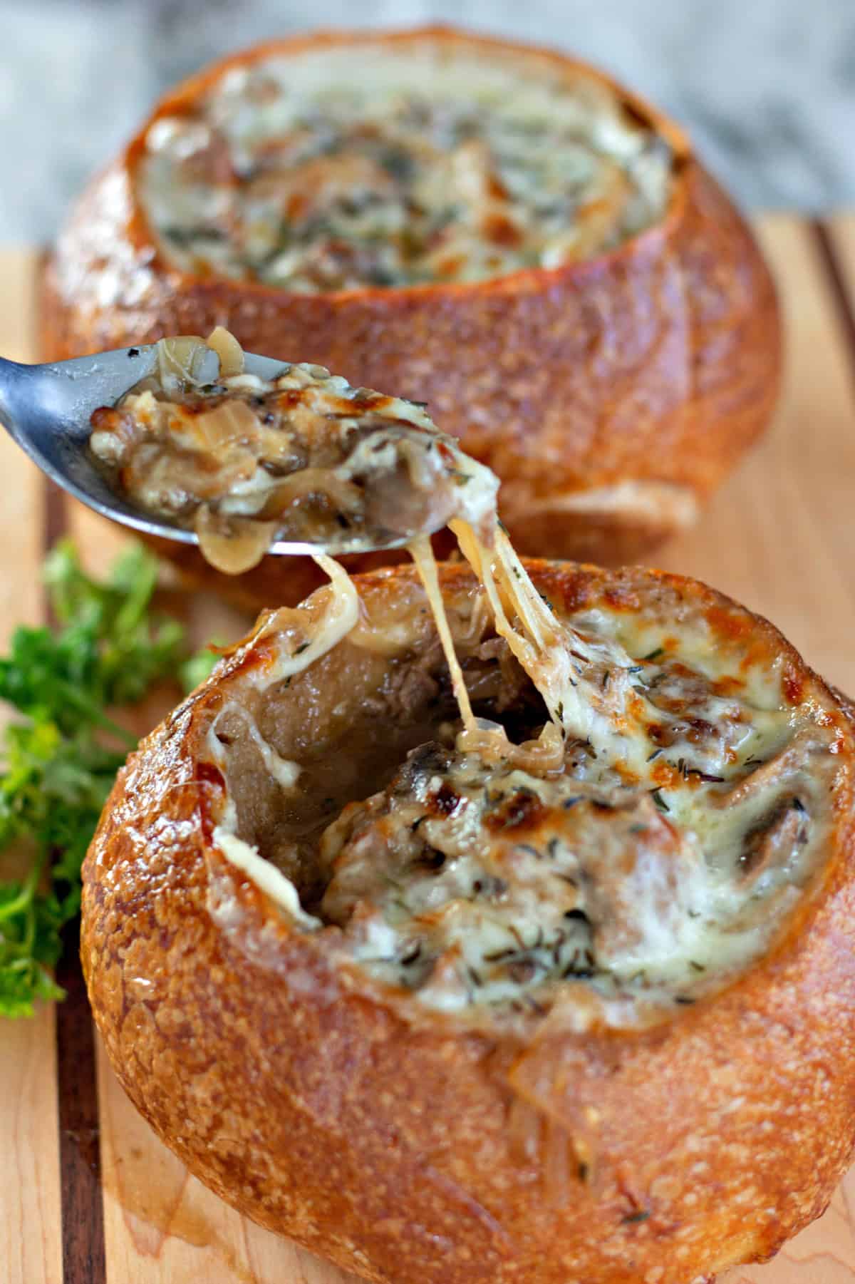 A top-down view of two bread bowls filled with Philly Cheesesteak Soup, topped with golden, bubbly provolone cheese and sprinkled with herbs. The removed bread tops rest beside them on a wooden cutting board, with a spoon and fresh parsley for garnish.