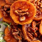 A close-up of glazed roasted sweet potato rounds topped with caramelized pecans and fresh herbs, glistening with syrupy sauce.