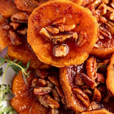 A close-up of glazed roasted sweet potato rounds topped with caramelized pecans and fresh herbs, glistening with syrupy sauce.