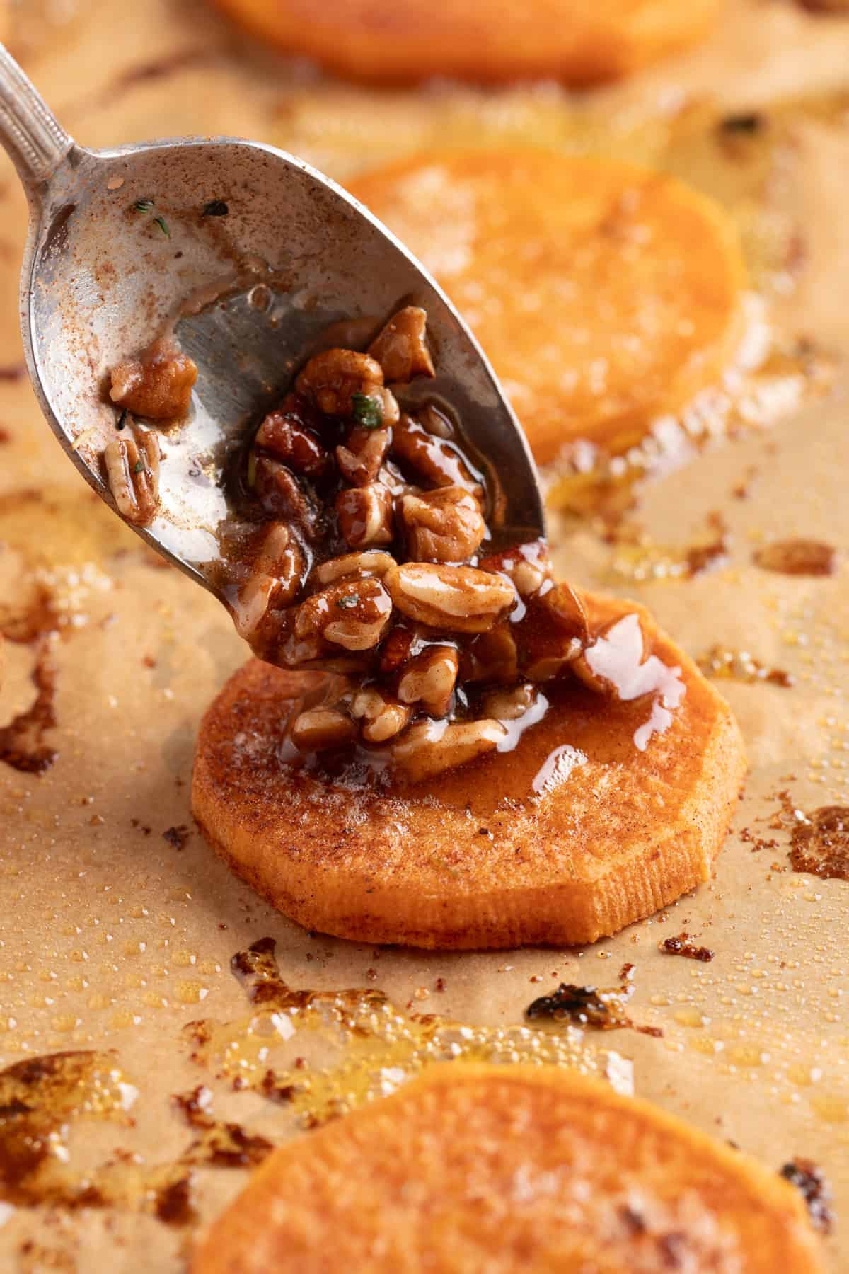 These simple Roasted Sweet Potatoes are a flavorful and versatile side dish that pairs perfectly with any meal. They’re caramelized on the outside, tender on the inside, and naturally sweet and buttery—my favorite way to enjoy sweet potatoes! A spoon drizzling the caramelized pecan topping over roasted sweet potato rounds.