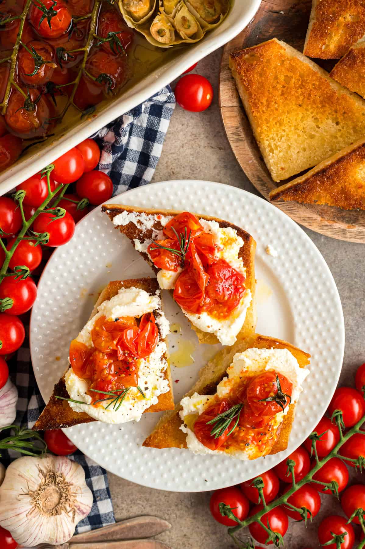 A beautifully arranged plate of ricotta toast topped with roasted cherry tomatoes, with fresh tomatoes and rustic kitchen decor in the background.