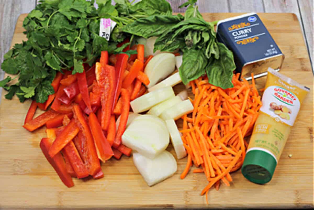 A wooden cutting board with fresh ingredients including sliced red bell peppers, onions, shredded carrots, cilantro, basil, curry powder, and a tube of minced ginger.