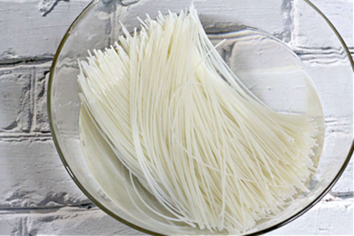 A glass bowl filled with uncooked rice noodles soaking in water, placed on a white brick-patterned surface.