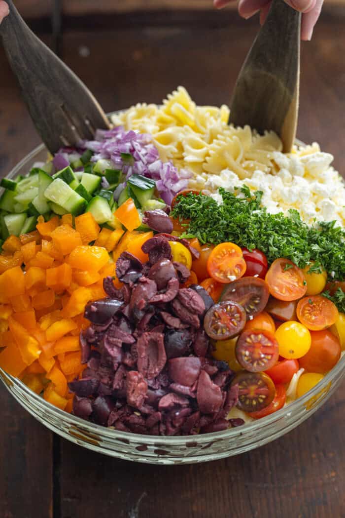 Wooden tongs stirring pasta, veggies, feta, and parsley in a bowl