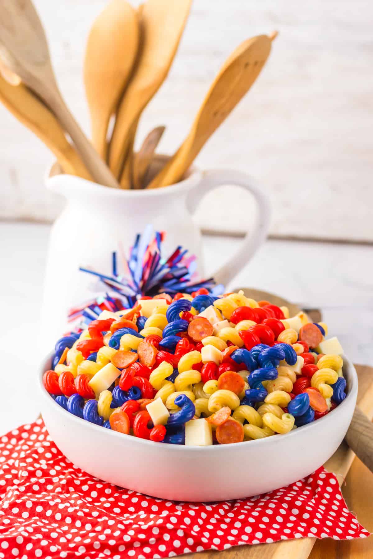 A patriotic pasta salad served in a white bowl, featuring red, yellow, and blue rotini pasta, mini pepperoni slices, and cubes of cheese. The bowl sits on a red polka dot napkin with a festive pom-pom and a white ceramic pitcher holding wooden utensils in the background.