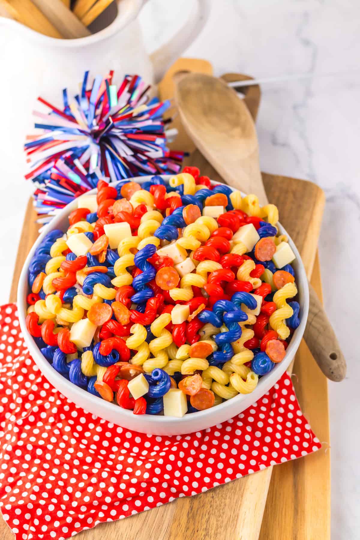 Patriotic pasta salad in a white bowl, filled with red, yellow, and blue rotini, cheese cubes, and pepperoni, set on a red polka dot napkin with a festive pom-pom decoration nearby.