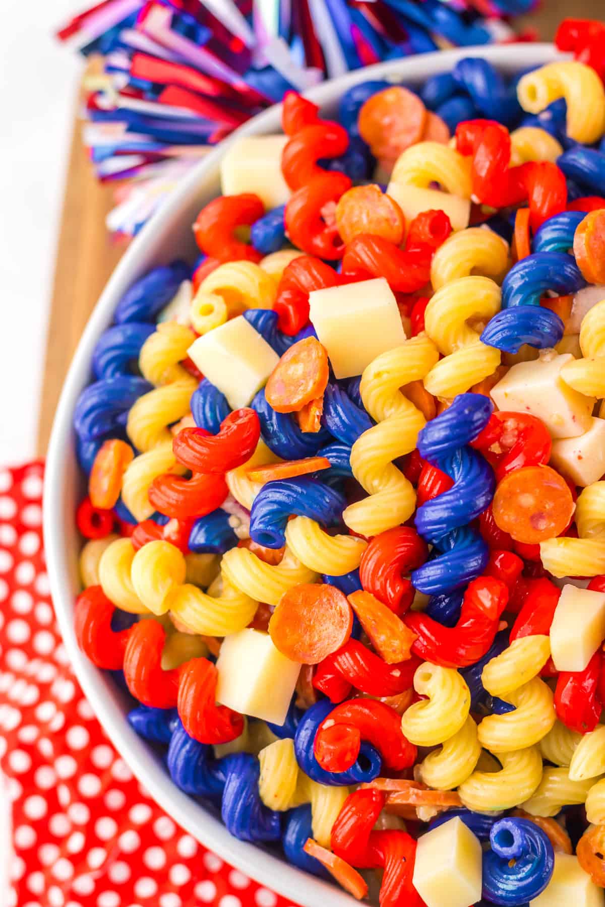 This 4th of July Pasta Salad is the ultimate festive side dish for your summer cookout! Top-down close-up of red, yellow, and blue rotini pasta salad, showing cheese cubes and slices of mini pepperoni nestled among the colorful pasta spirals.