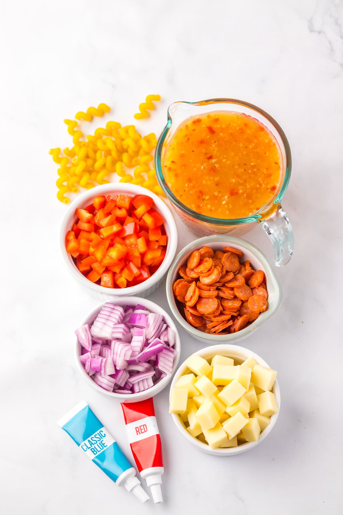 Flat lay of pasta salad ingredients, including uncooked cavatappi pasta, diced red bell pepper, mini pepperoni slices, chopped red onion, cubed cheese, Italian dressing in a glass measuring cup, and red and blue gel food coloring tubes.