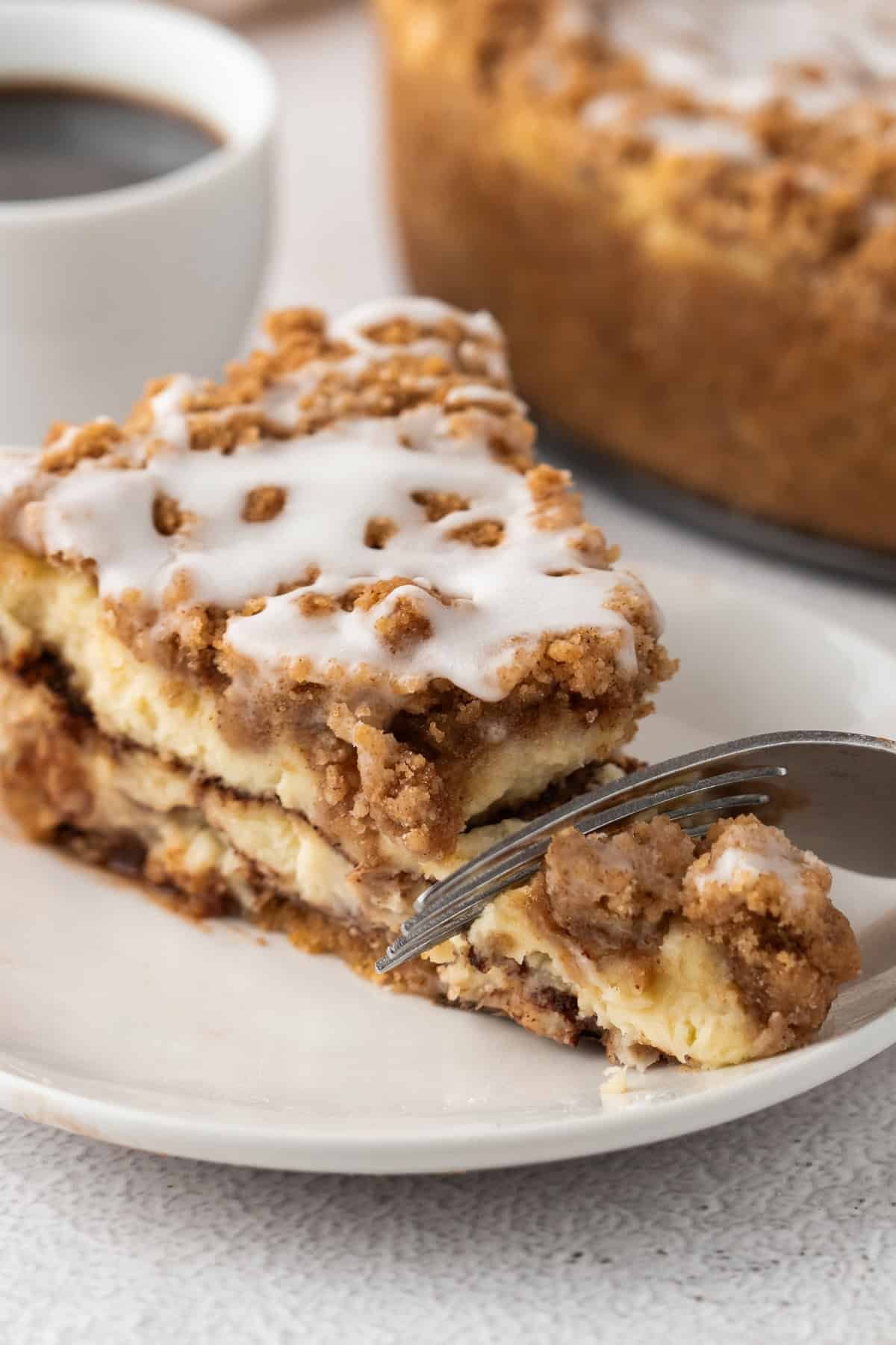 A close-up of a fork cutting into a slice of coffee cake cheesecake, highlighting the soft layers of cheesecake and cinnamon crumb topping with a glossy icing finish.