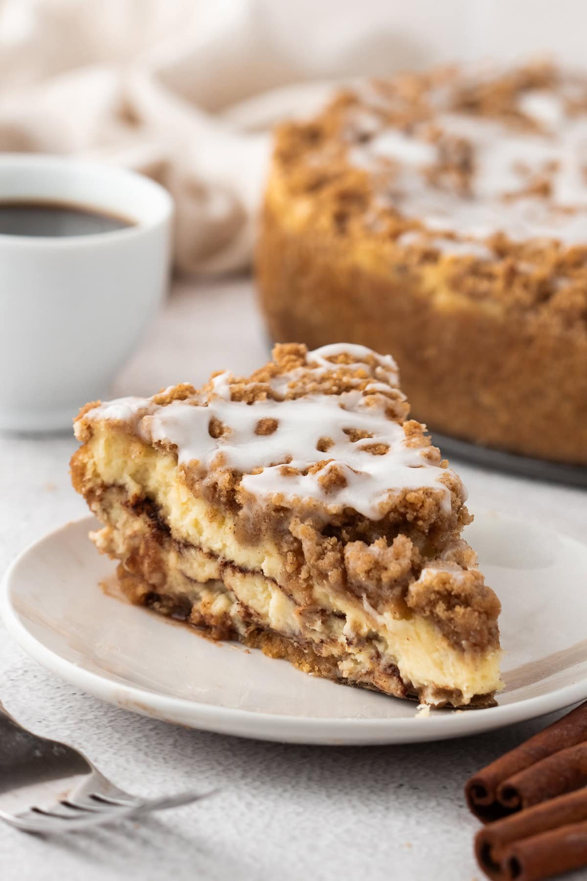 Coffee Cake Cheesecake is a delicious layered dessert that’s the perfect mash-up of cheesecake and coffee cake. Its lovely balanced flavors and textures make it addictive. A plated slice of coffee cake cheesecake with a cup of coffee in the background. The cake is topped with a thick cinnamon streusel and icing, and the whole cheesecake is visible behind the slice.
