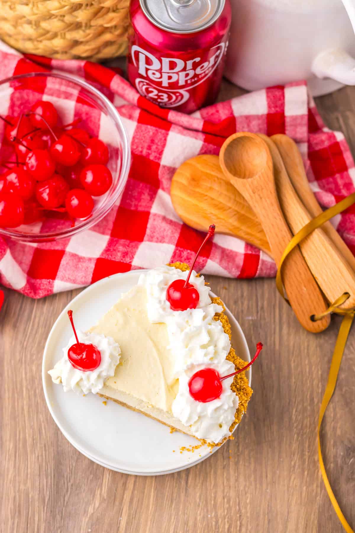 Overhead shot of a single slice of Dr Pepper pie with whipped cream and cherries, placed on a white plate next to a bowl of cherries and a can of Dr Pepper.