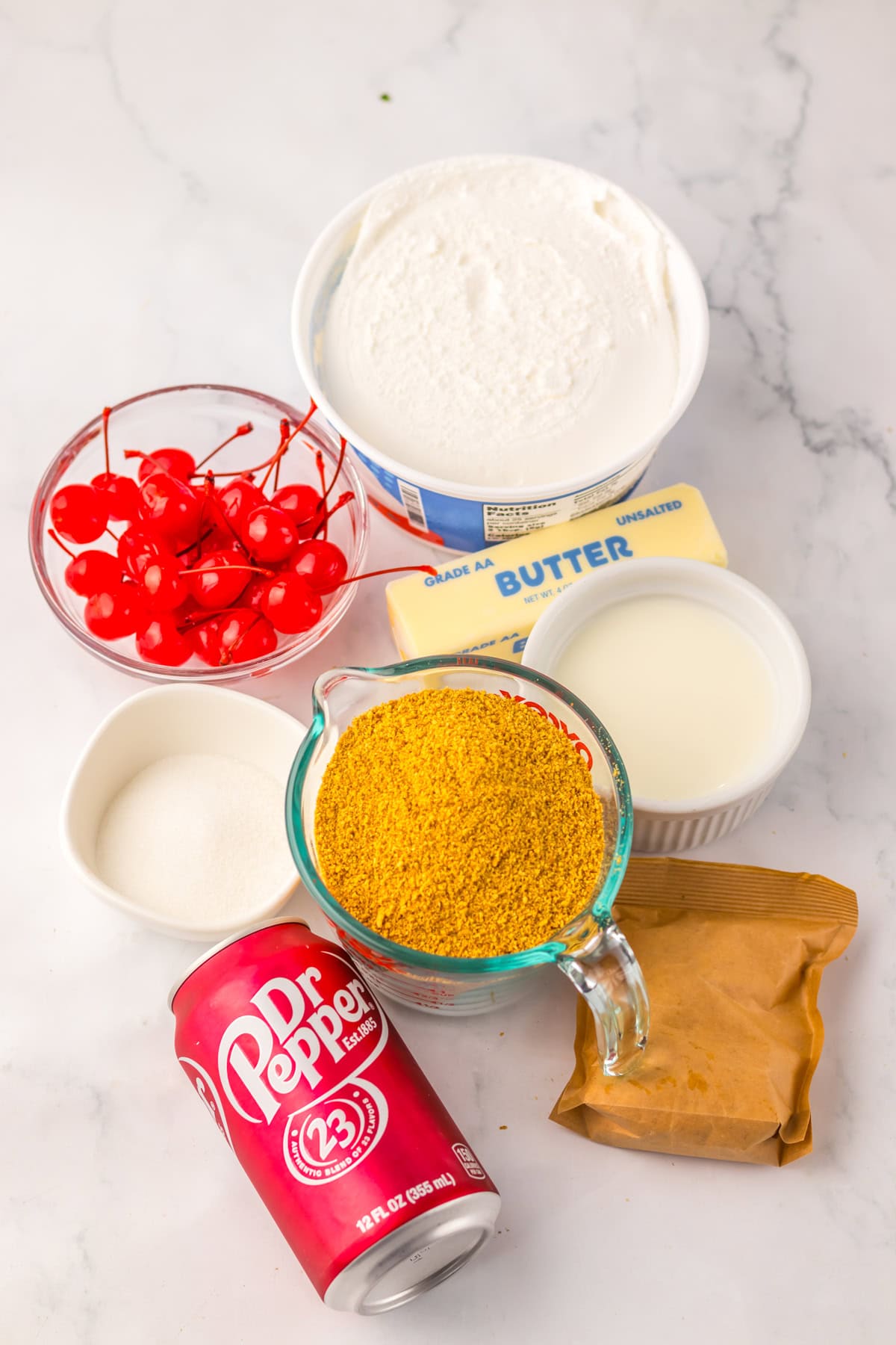 Flat lay of ingredients for Dr Pepper pie, including a can of Dr Pepper, maraschino cherries, whipped topping, unsalted butter, milk, granulated sugar, graham cracker crumbs, and an instant pudding mix packet on a marble surface.