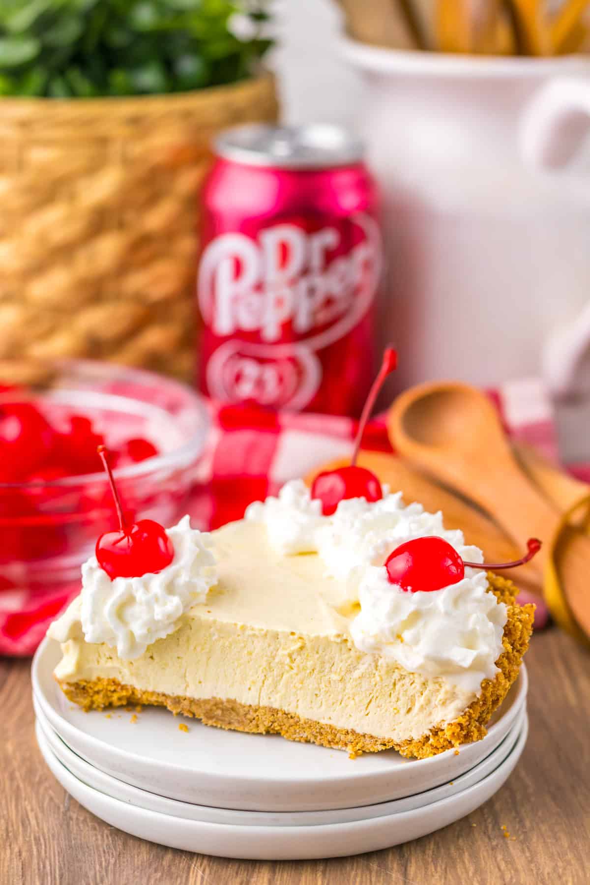 This Dr Pepper Pie is a rich, creamy frozen dessert that puts a fun twist on a classic soda favorite. With its smooth texture and signature cherry-cola flavor, it’s incredibly easy to make and the perfect no-bake treat for hot days or last-minute gatherings! Side view of a slice of Dr Pepper pie with a graham cracker crust, whipped cream, and cherries, styled with kitchen tools and red checkered cloth.