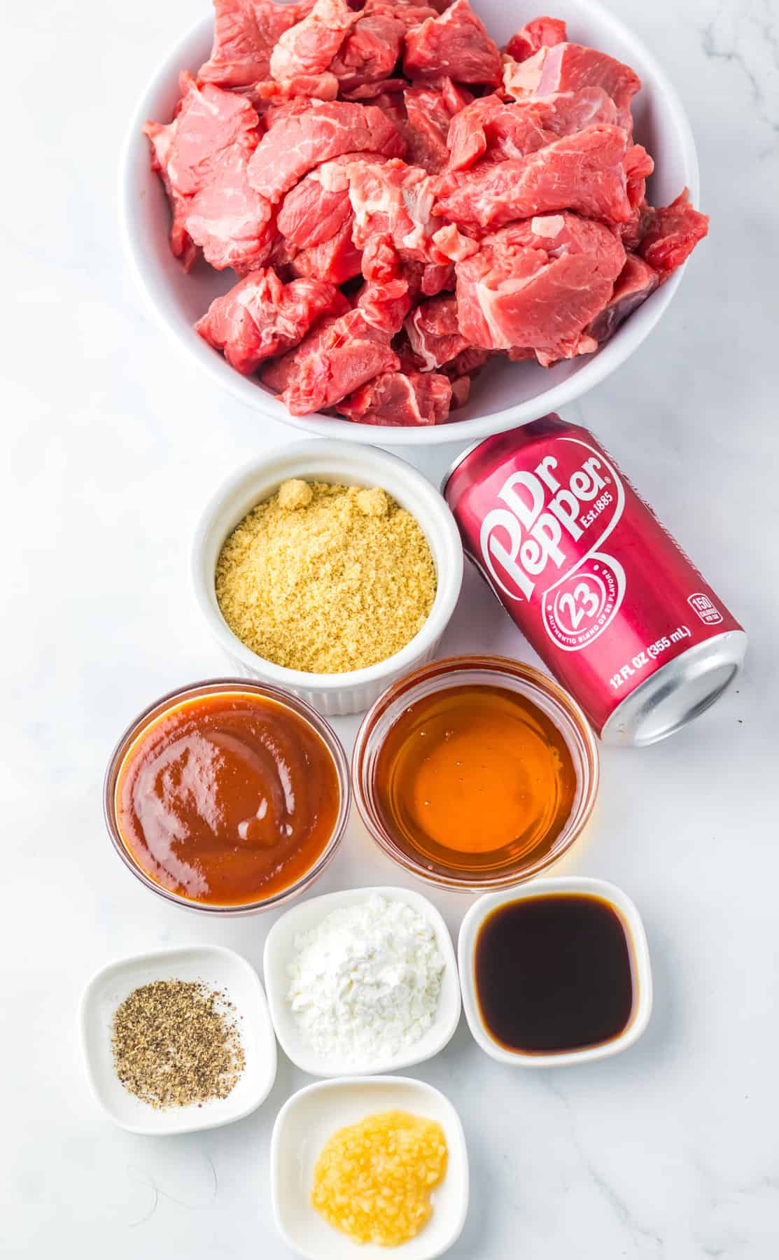 Overhead view of ingredients for Dr Pepper steak bites, including a bowl of raw beef cubes, a can of Dr Pepper, brown sugar, barbecue sauce, honey, soy sauce, garlic, cornstarch, pepper, and seasoning in small bowls arranged on a white surface.