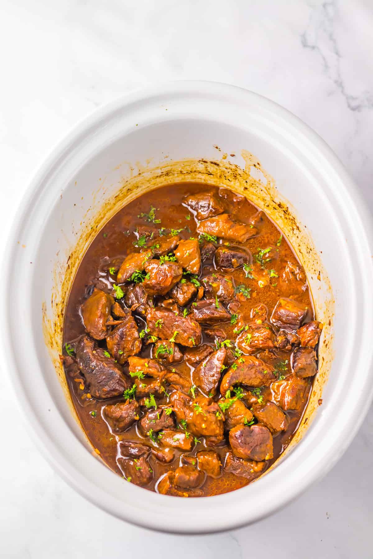 Slow cooker filled with cooked Dr Pepper steak bites in a thick, rich sauce, garnished with chopped herbs.