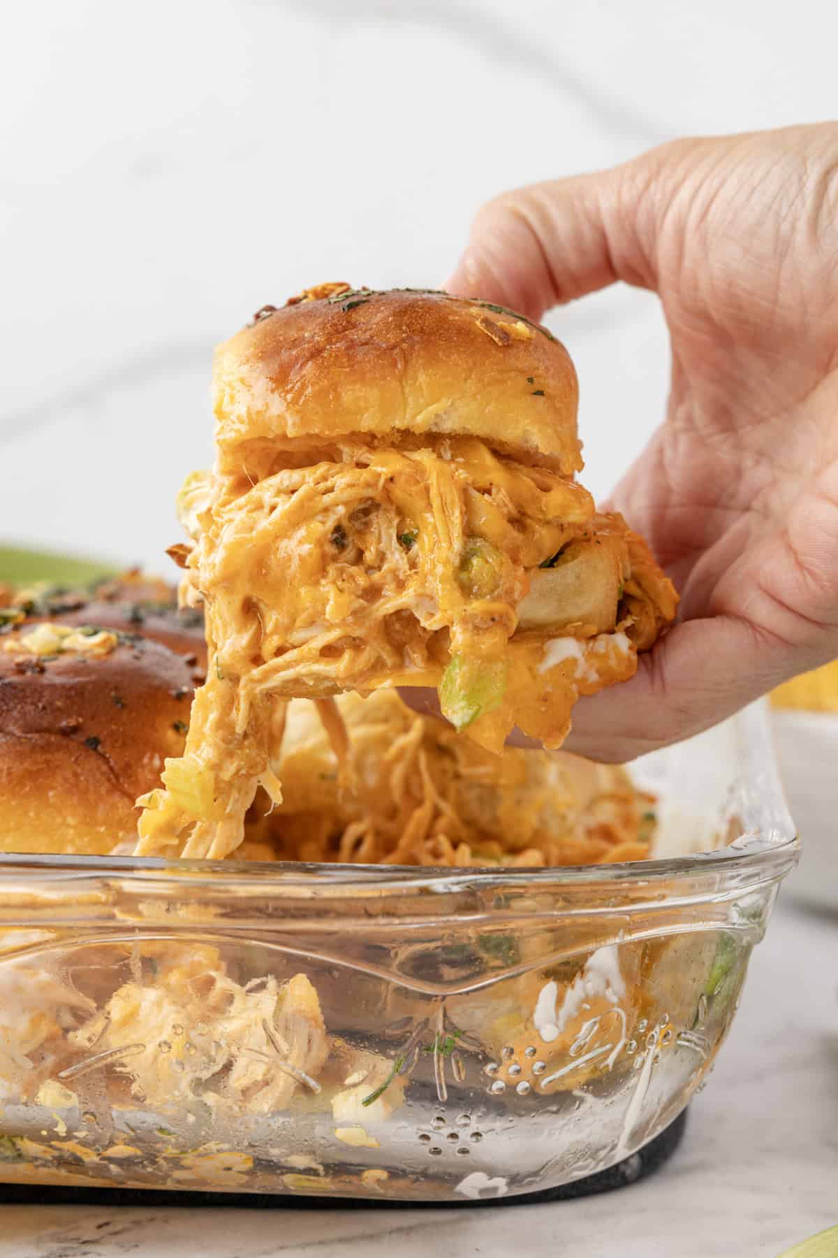 A hand pulling a buffalo chicken slider from a baking dish