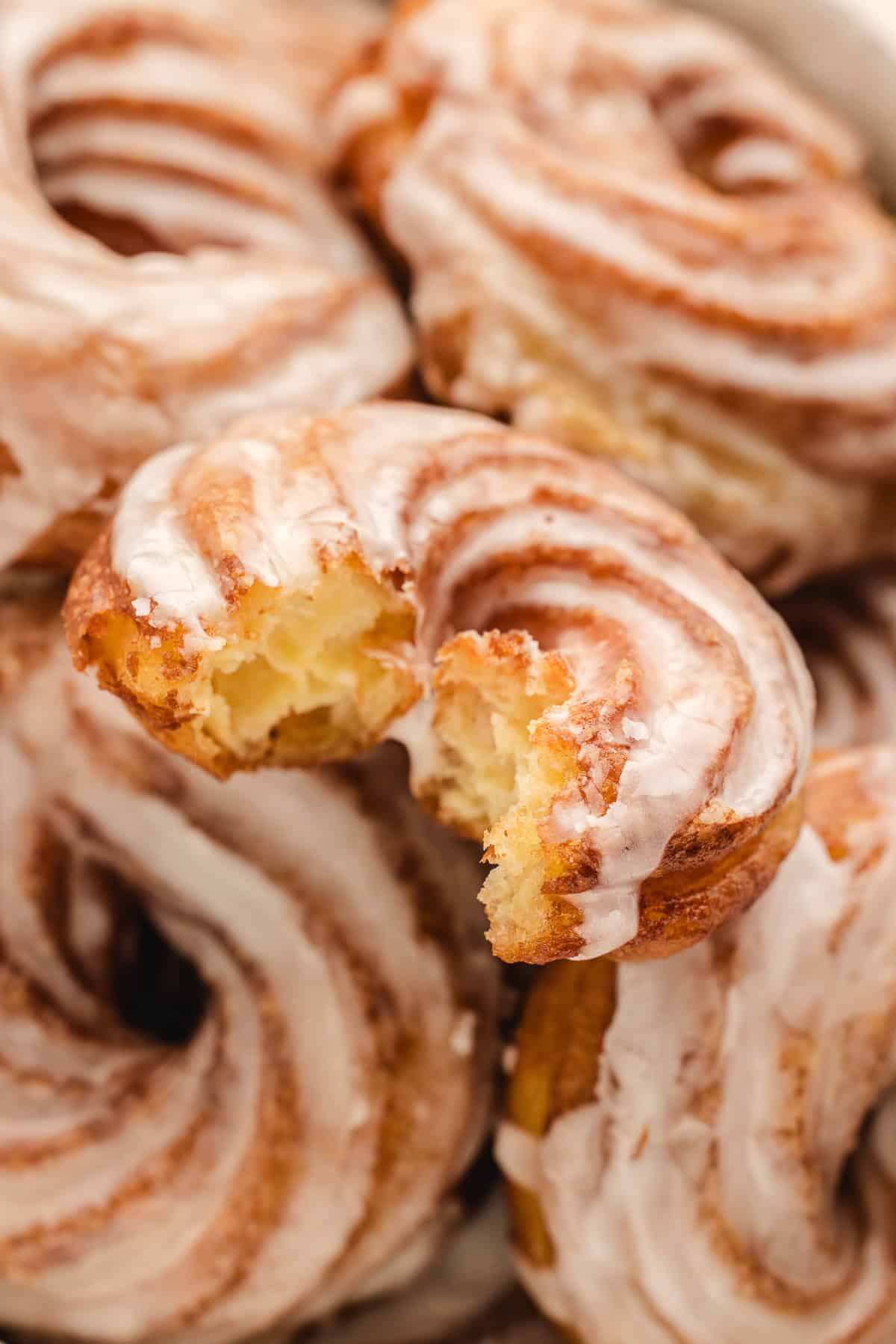 A pile of glazed French cruller donuts, one with a bite missing