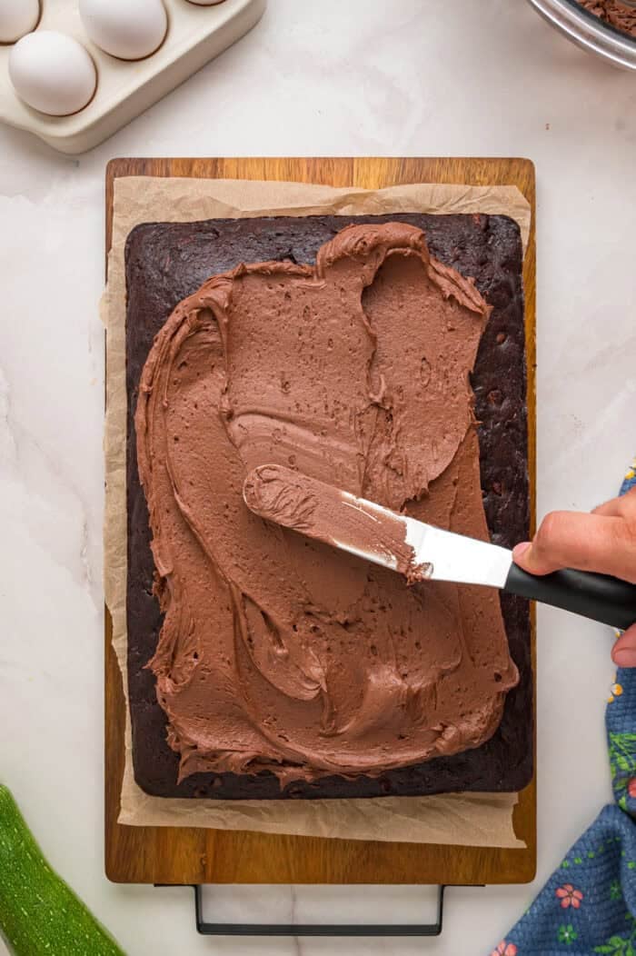 This Chocolate Zucchini Cake is rich, moist, and topped with an irresistible chocolate buttercream! It's a must-bake every summer! Spreading chocolate buttercream on chocolate zucchini cake