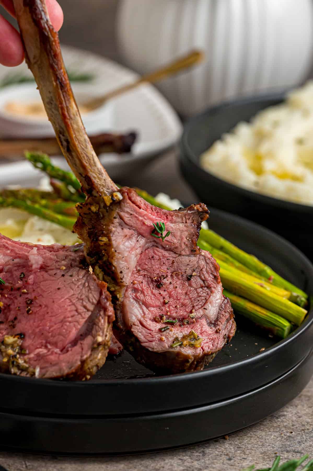 A hand holding a lamb chop vertically on a black plate