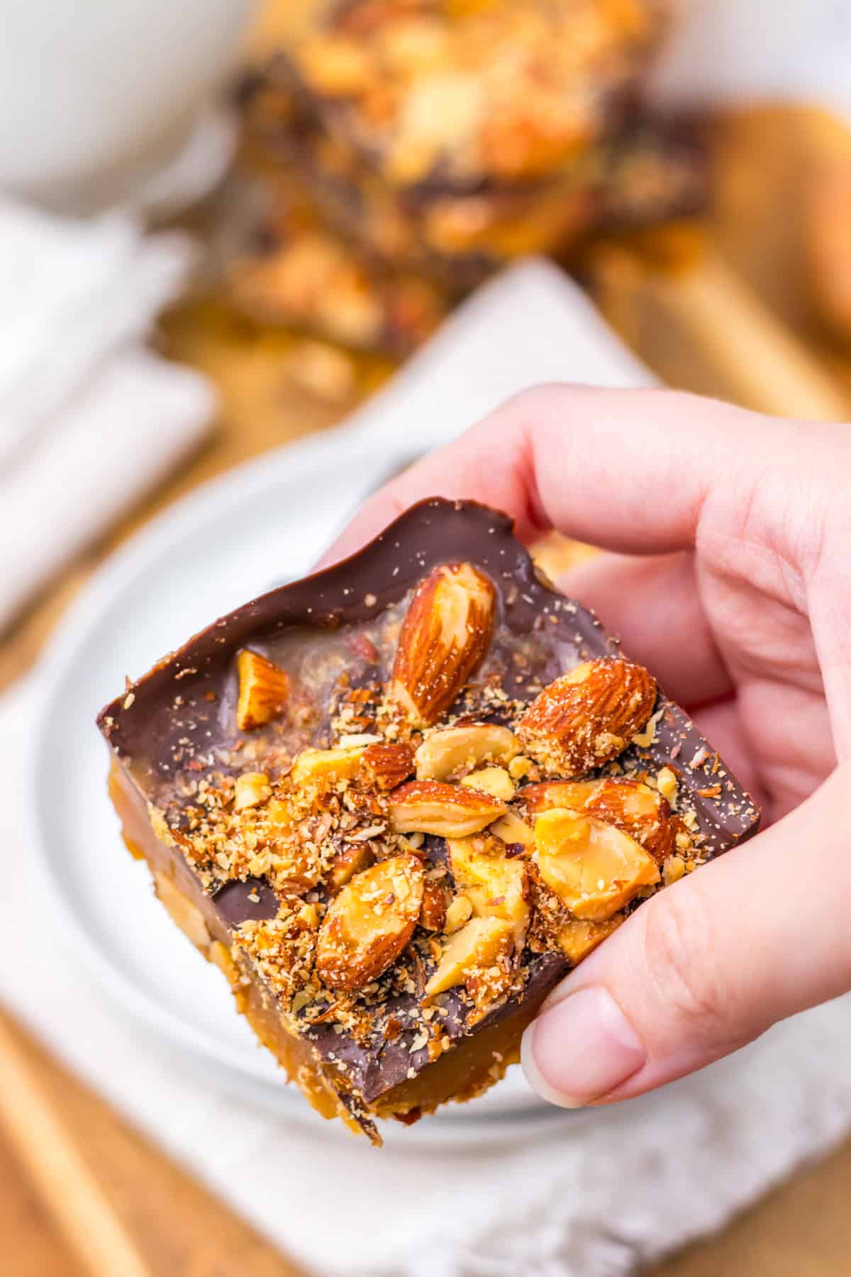 With just four ingredients, this Almond Roca Recipe features layers of almonds, toffee, and chocolate. Buttery, crunchy, and easy to make! A hand holding a piece of almond roca