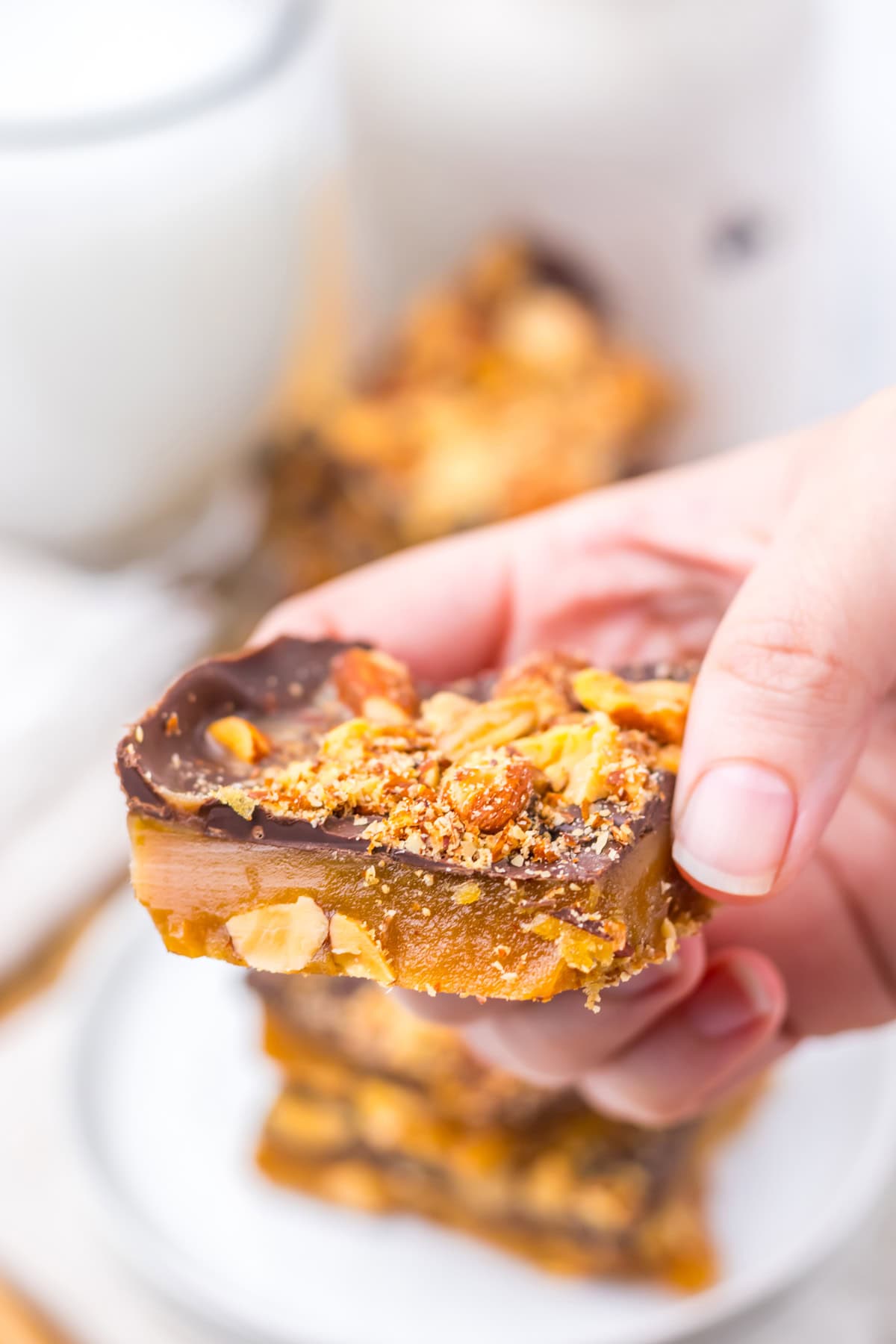 With just four ingredients, this Almond Roca Recipe features layers of almonds, toffee, and chocolate. Buttery, crunchy, and easy to make! A hand holding a piece of homemade almond roca