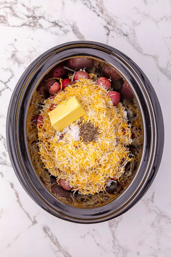 Cheddar cheese, butter, and spices on top of bacon and potatoes in a crockpot