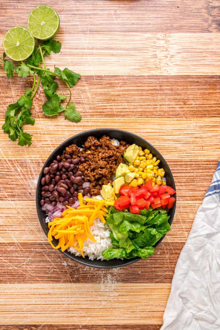 Elevate Taco Tuesday with this easy Taco Bowl recipe. With seasoned taco meat, rice and cilantro lime crema, it's an easy weeknight dinner. Ground beef, corn, tomatoes, lettuce, and cheese on a bowl of lettuce