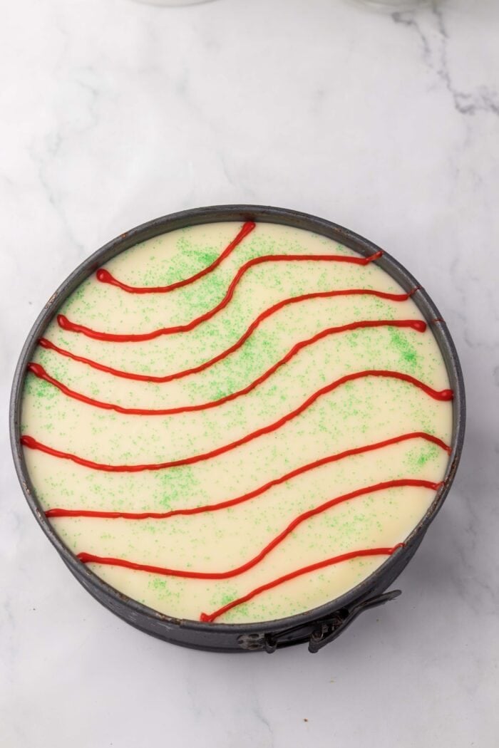 With no baking required, this Little Debbie Christmas Tree Cheesecake is a fun, festive, and nostalgic Christmas treat. Red icing drizzled over white ganache in a springform pan
