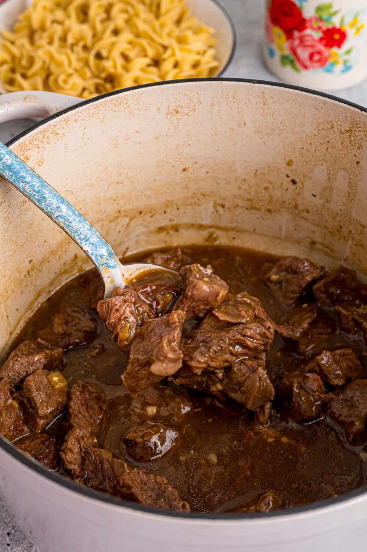 Tender chunks of beef are cooked in a rich, savory gravy and served over egg noodles in this easy Beef and Noodles recipe. A spoon holding the beef from beef and noodles over a Dutch oven