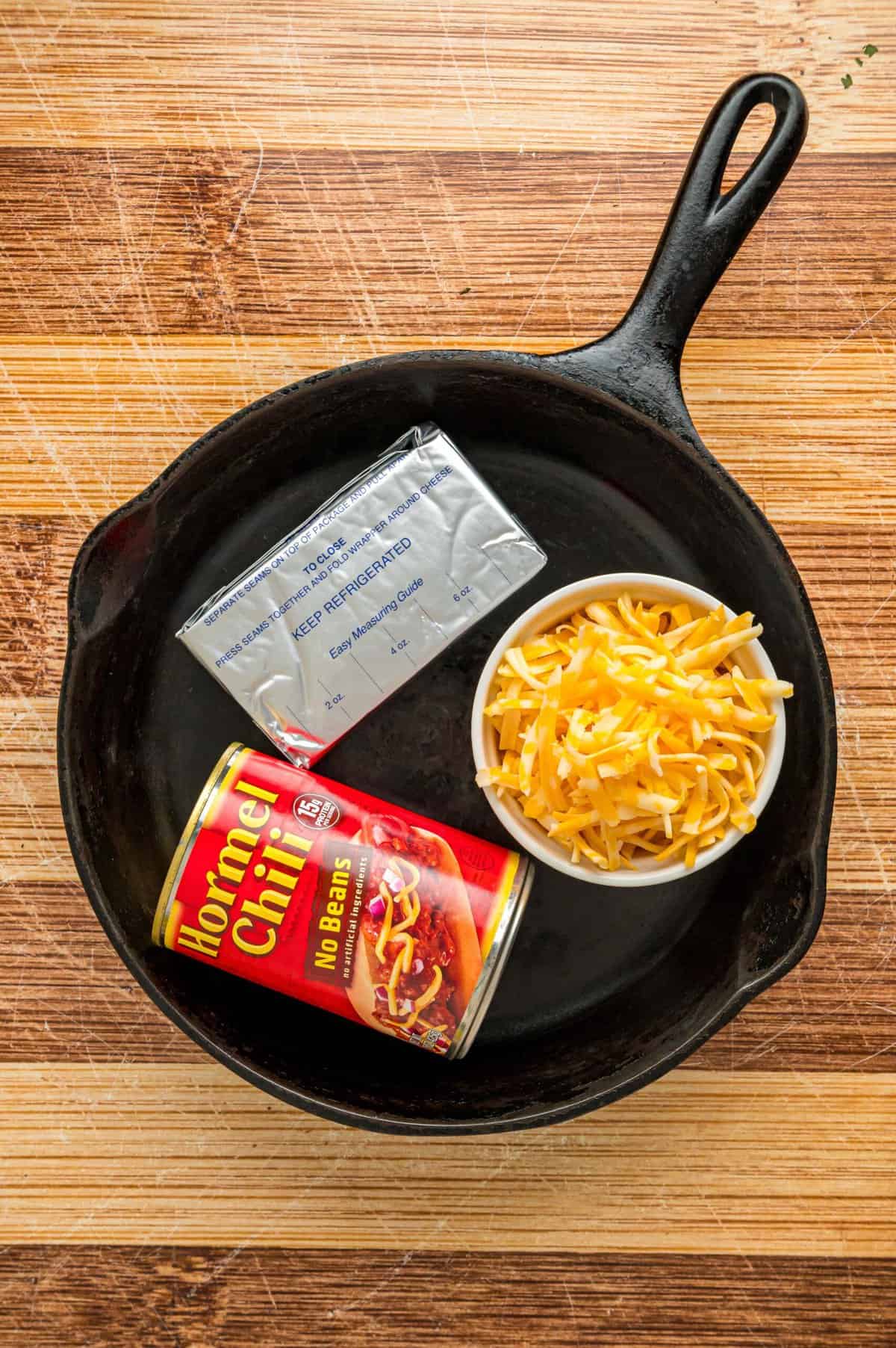A can of chili, shredded cheese, and cream cheese in a cast iron skillet