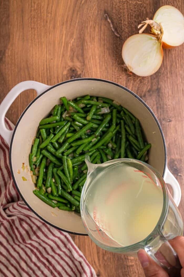 Fresh green beans are slow-cooked until tender with garlic, onion, bacon, and some Cajun seasoning in this Southern Green Beans recipe. Pouring broth over fresh green beans in a pot