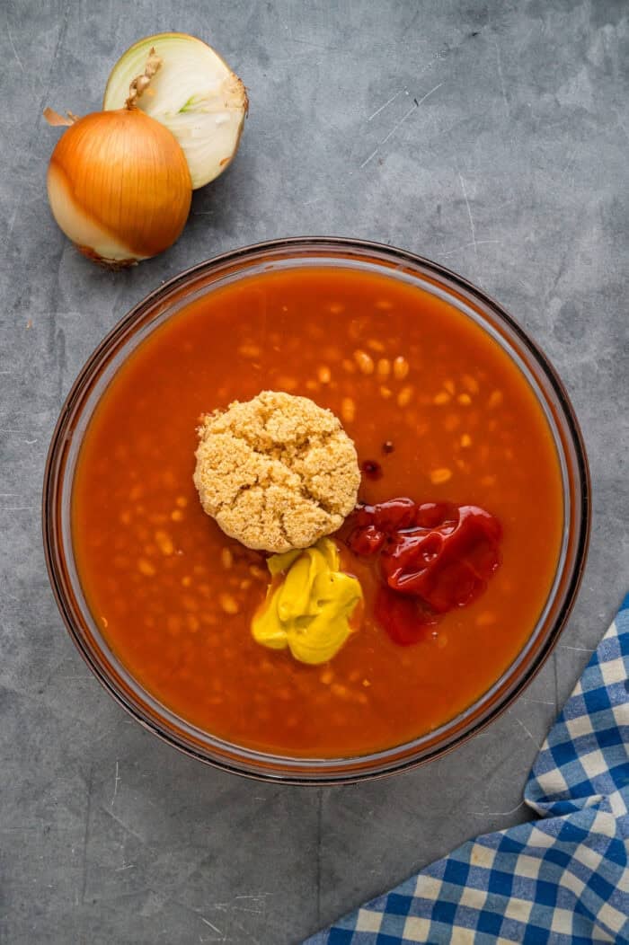 This easy Baked Beans recipe begins with pork and beans to keep things simple and salty bacon to add a final touch. Great for BBQs! Pork and beans, brown sugar, ketchup, and mustard in a bowl