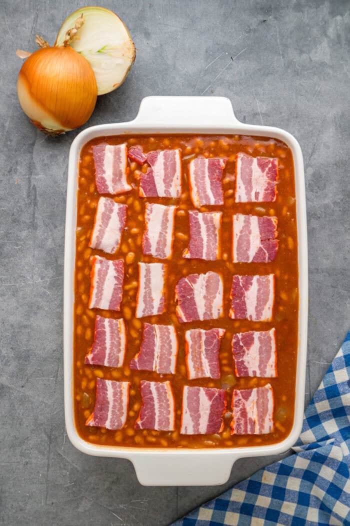 This easy Baked Beans recipe begins with pork and beans to keep things simple and salty bacon to add a final touch. Great for BBQs! Bacon slices on top of beans in a baking dish