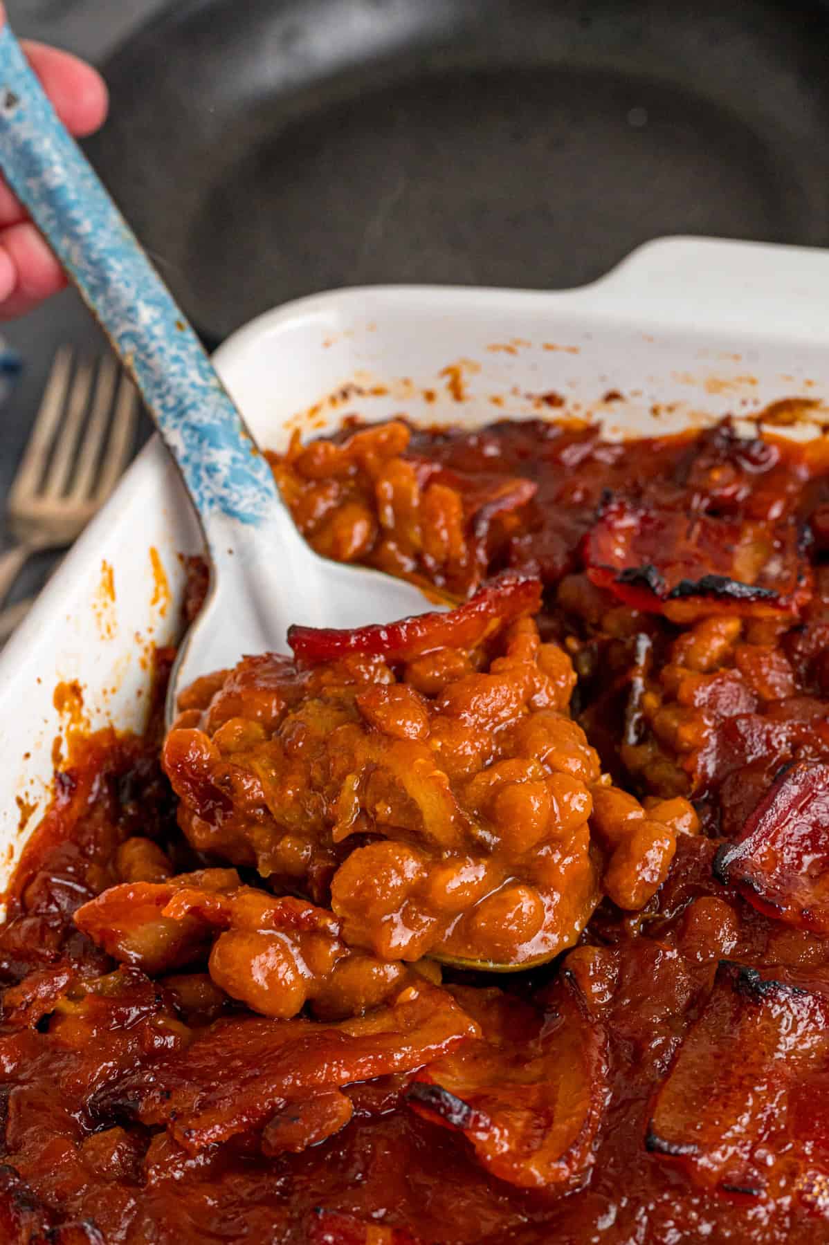 This easy Baked Beans recipe begins with pork and beans to keep things simple and salty bacon to add a final touch. Great for BBQs! A serving spoon in a baking dish of baked beans