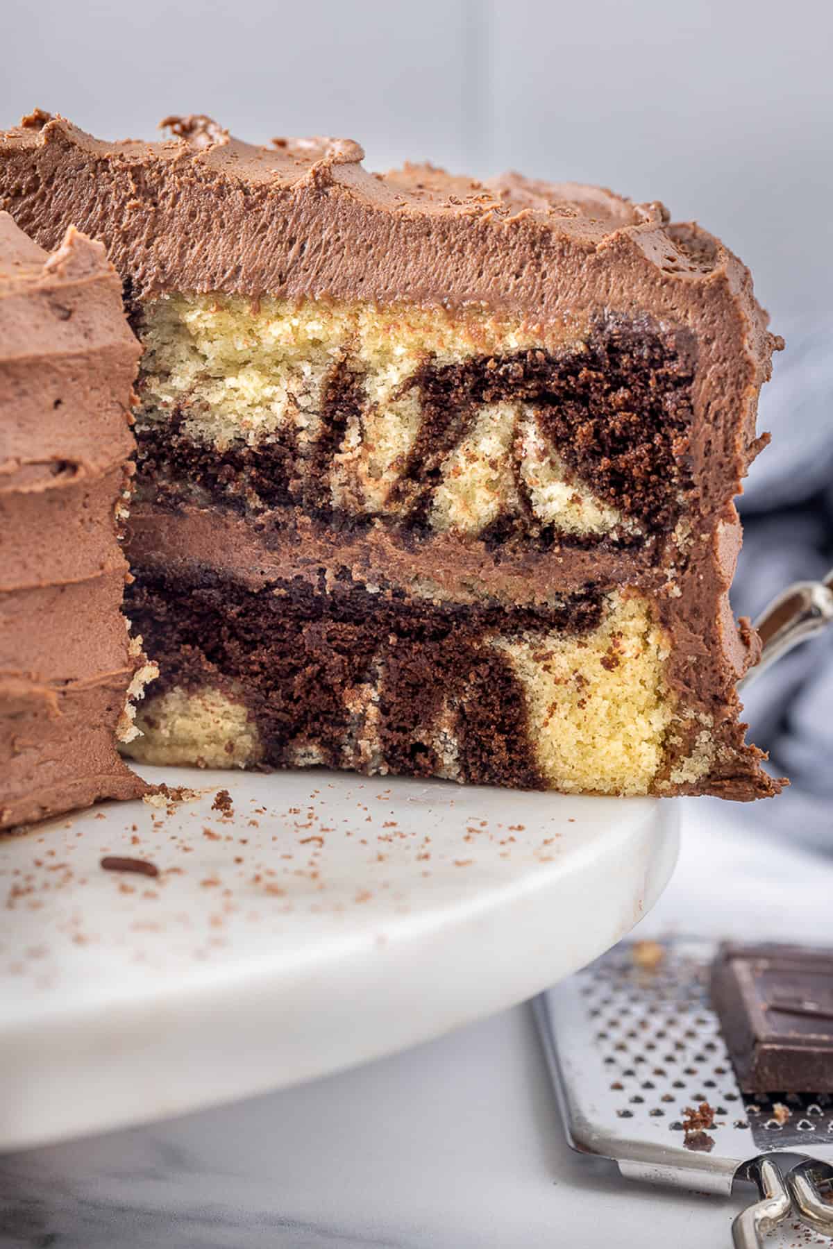 Chocolate and vanilla cake batter are swirled together, baked, and then layered with chocolate buttercream to create this Marble Cake recipe. Marble cake on a cake stand with one slice missing to show the layers