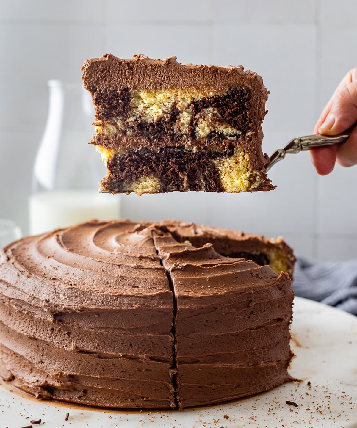 Chocolate and vanilla cake batter are swirled together, baked, and then layered with chocolate buttercream to create this Marble Cake recipe. A slice of marble cake on a spatula above the cake